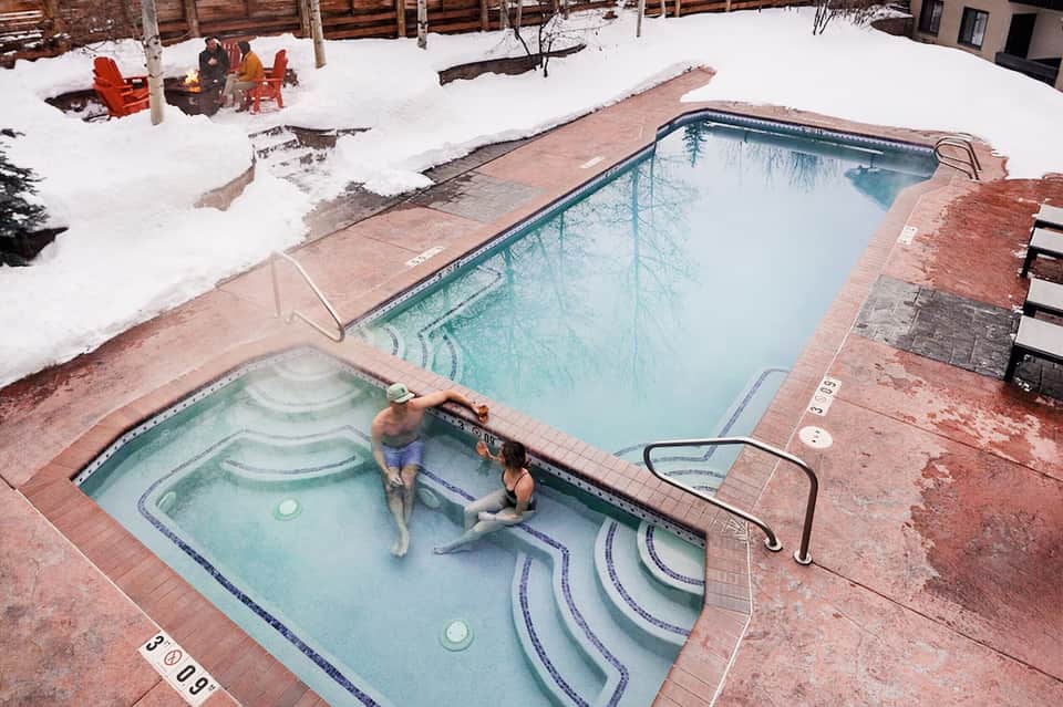 Guests enjoying hot tub in snow-covered resort courtyard with heated pool in background
