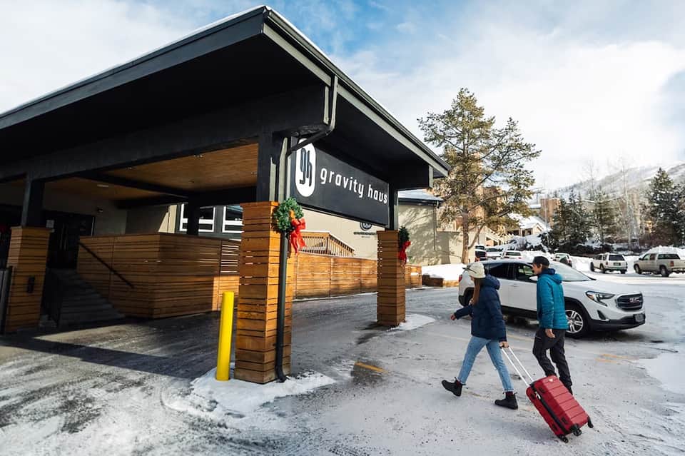 Gravity House mountain lodge exterior with wooden slat details and guests arriving with luggage in snowy setting