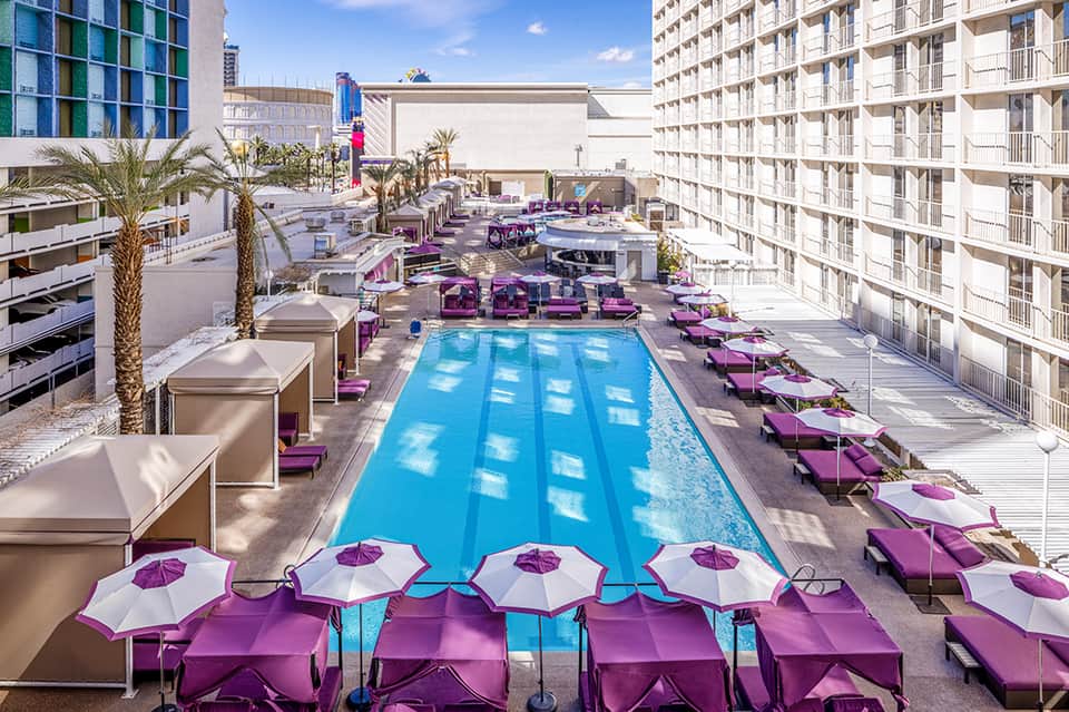 Resort pool deck with purple loungers and umbrellas, surrounded by modern high-rise buildings under clear sky