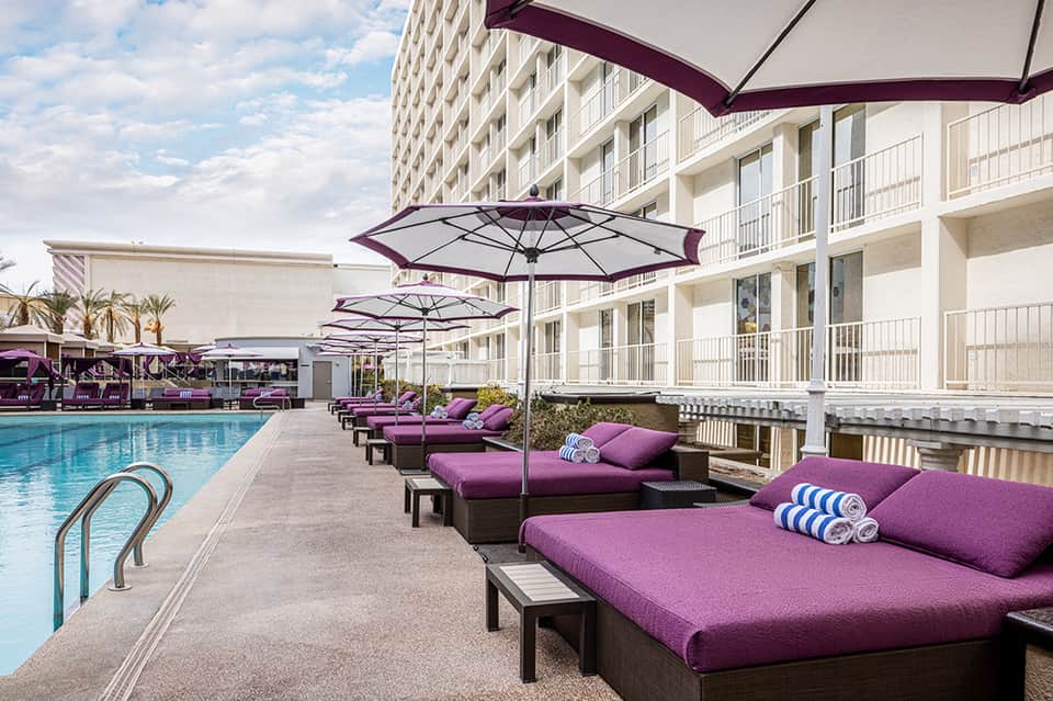 Resort poolside with purple lounge chairs, umbrellas, and multi-story hotel building in background