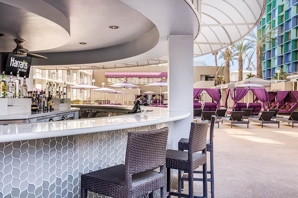 Modern hotel bar with curved white counter, rattan chairs, and purple cabana seating area overlooking palm trees