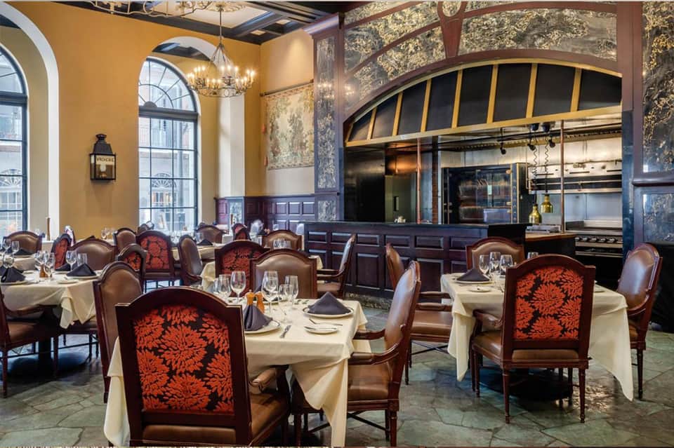 Upscale fine dining restaurant with gold walls, arched windows, open kitchen, and tables set with cream linens and red patterned chairs