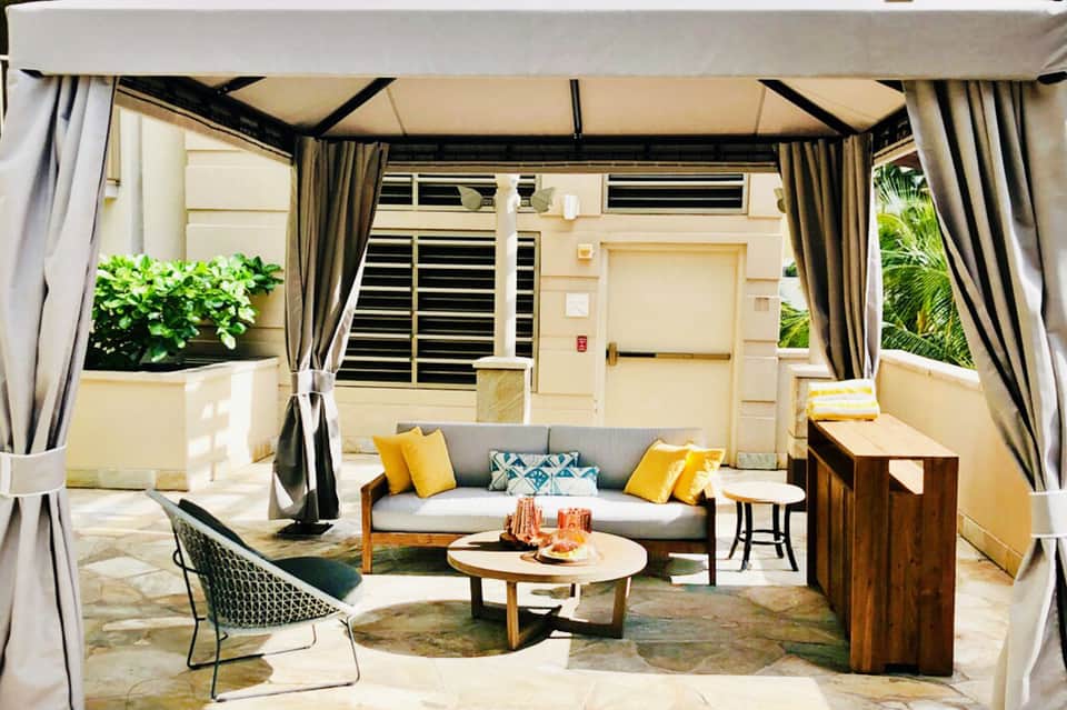 Covered patio lounge with gray sofa, yellow pillows, wooden tables, and draped curtains overlooking a building entrance