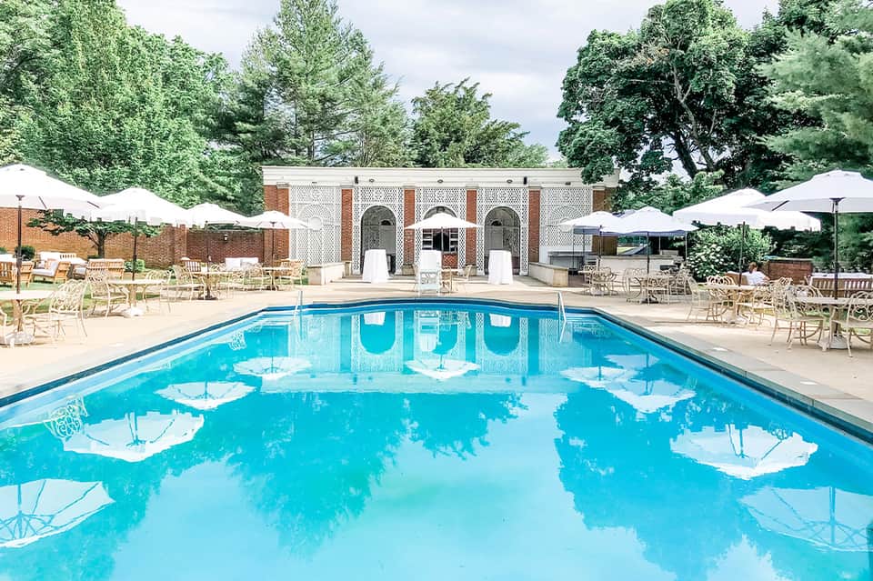 Resort pool with blue water, white umbrellas, brick building with arched architecture, lounge chairs