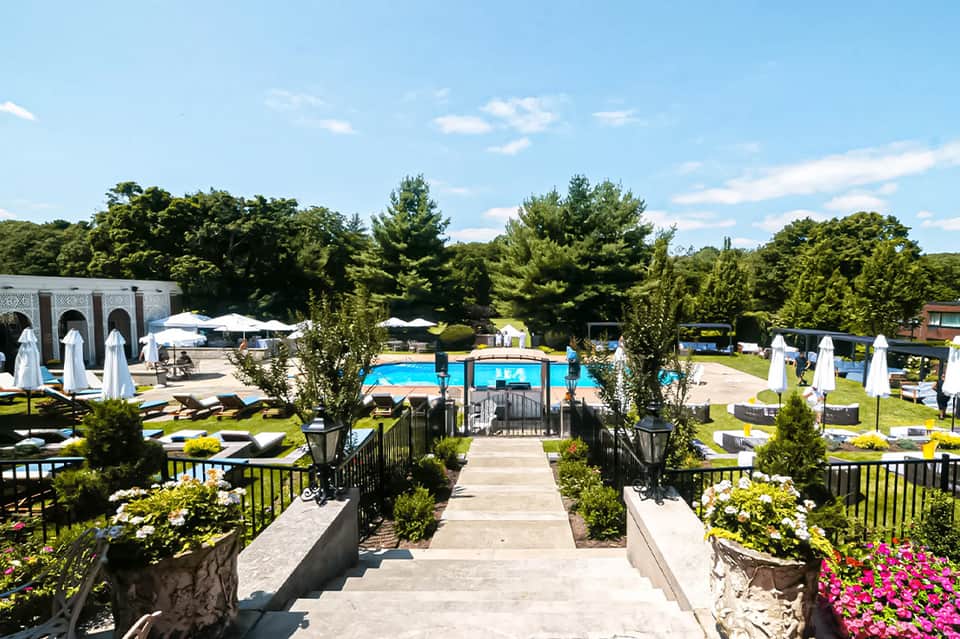 Resort pool area with blue water, white umbrellas, manicured gardens, and flowering plants along a concrete pathway