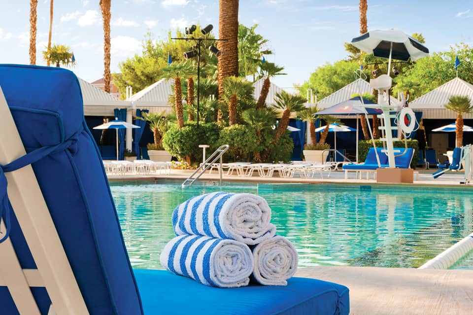 Resort pool with blue lounge chairs, rolled towels, palm trees, and white umbrellas under bright sunshine