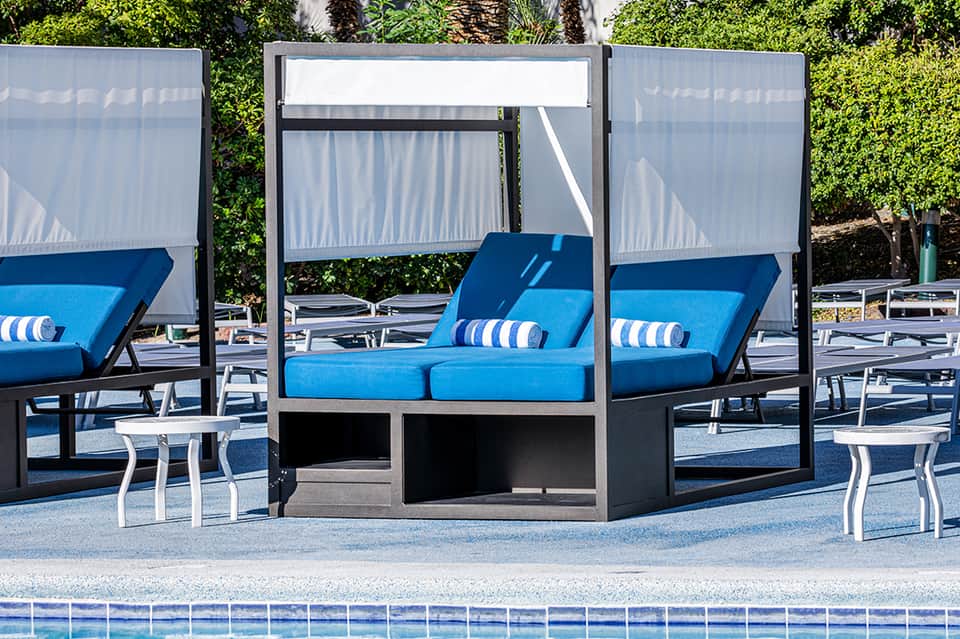 Poolside lounge area with blue cushioned daybed, white retractable shade, and green landscaping