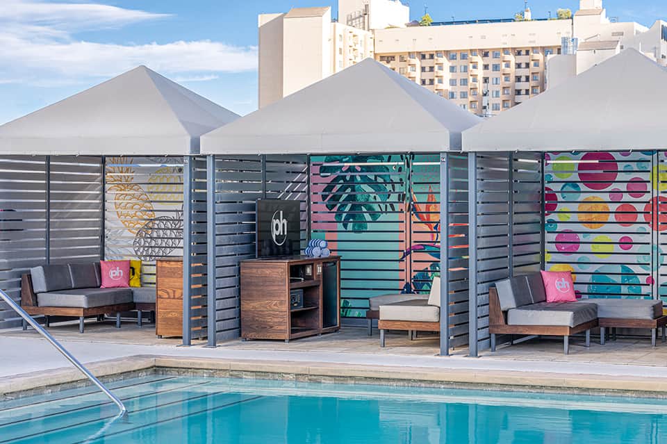 Modern pool deck with white tent canopies, colorful graphic privacy screens, wooden lounge furniture, and high-rise buildings in background