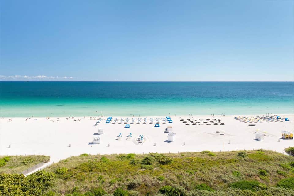 Aerial view of a crowded beach with turquoise water, white sand, and blue beach umbrellas