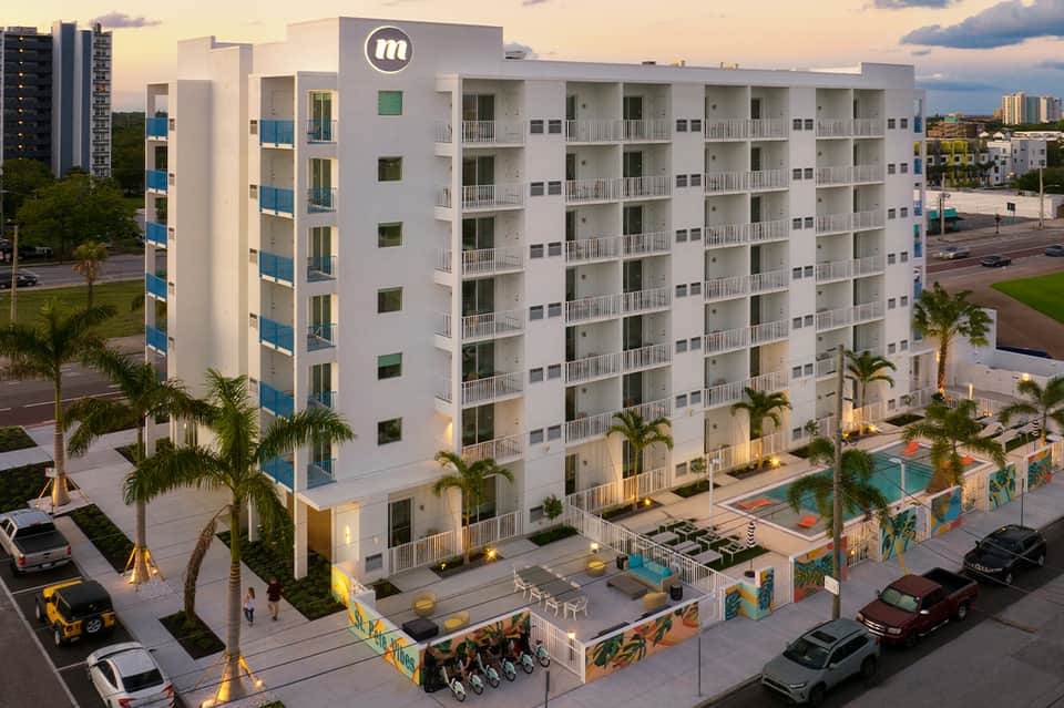 Modern white apartment building with blue accents, ground-level pool, colorful murals, and palm trees at dusk