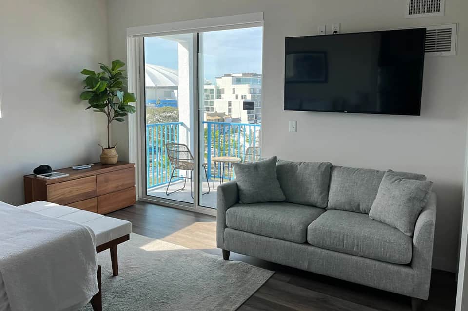 Modern hotel room with gray sofa, ocean view balcony, wall-mounted TV, wooden dresser, and potted plant