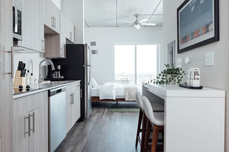 Compact studio hotel room with kitchenette, white bedding, desk area, and floor-to-ceiling windows.