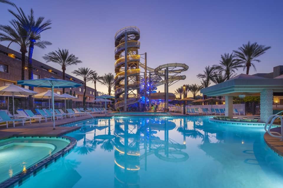 Illuminated resort pool complex at dusk with water slides, palm trees, and lounge chairs