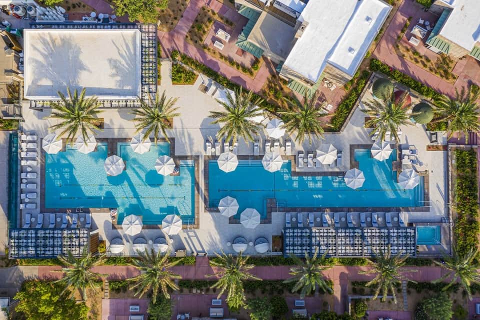 Aerial view of resort pools surrounded by palm trees, umbrellas, and sandy grounds