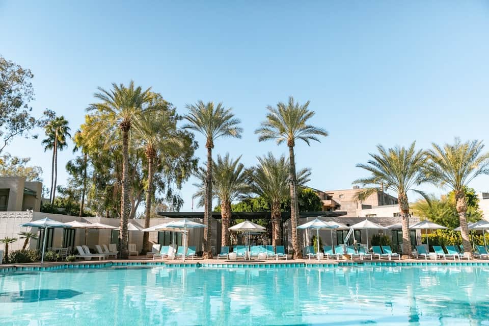 Resort pool with crystal-clear turquoise water, palm trees, and lounge chairs with white umbrellas under blue sky