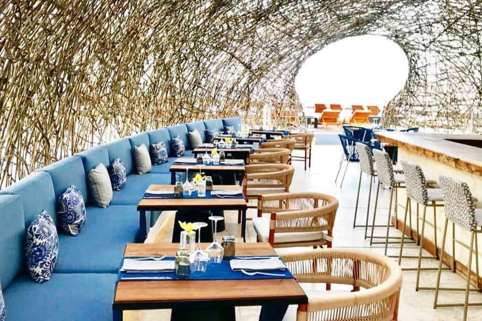 Modern restaurant terrace with blue banquette seating, woven chairs, and reed wall screening