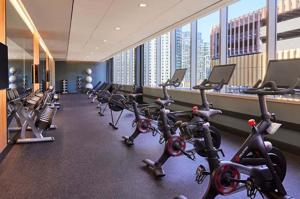Modern fitness center with stationary bikes, treadmills, and floor-to-ceiling windows overlooking city buildings