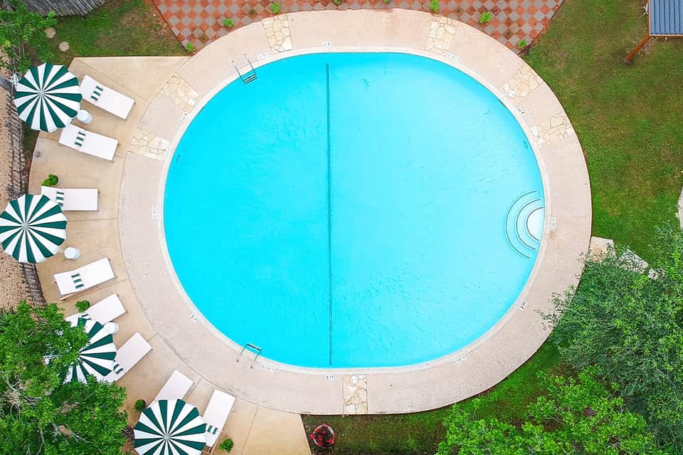Aerial view of circular turquoise swimming pool with beige deck and green umbrellas with lounge chairs