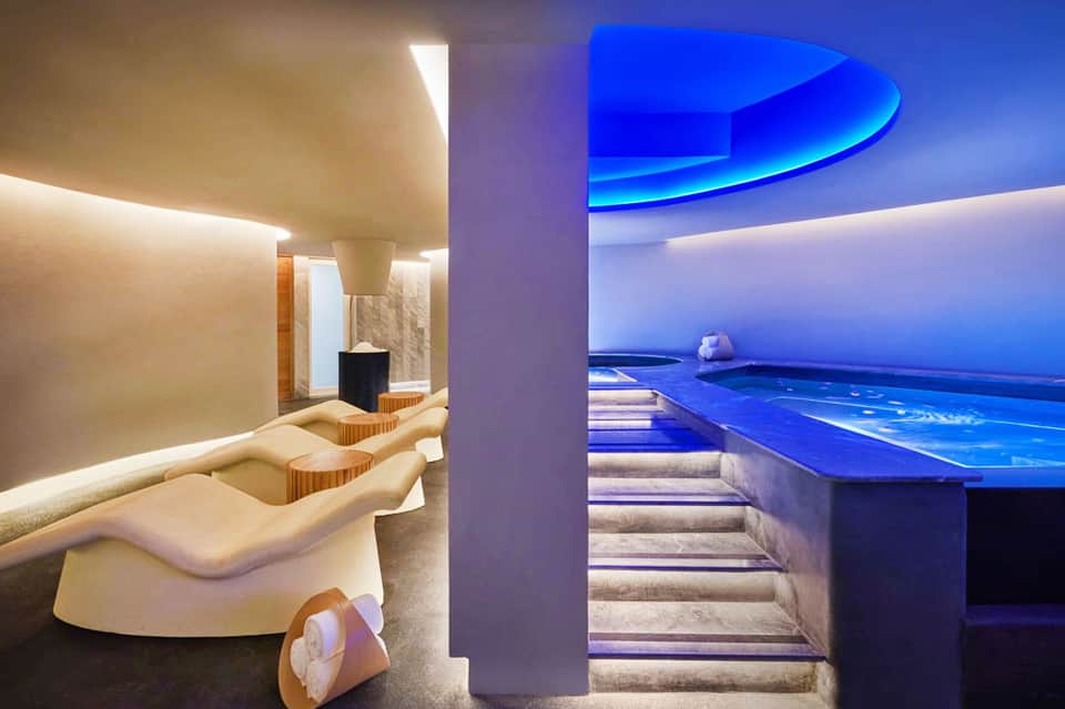 Modern spa interior with curved white seating, warm lighting, and blue infinity pool overlooking ocean