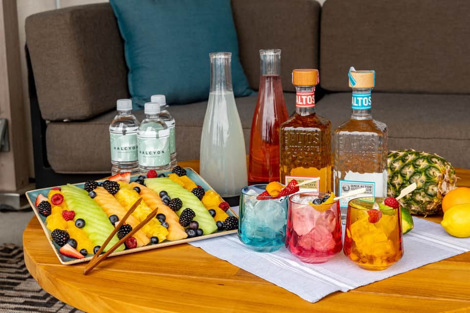 Colorful cocktail setup with fresh fruit platter, spirits bottles, and mixed drinks on wooden table