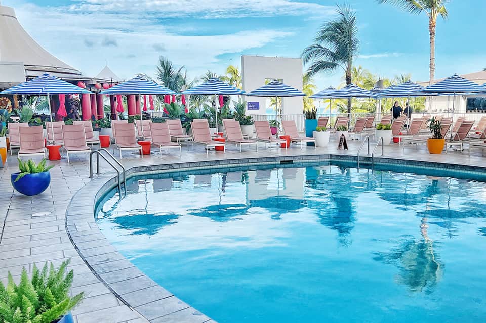 Resort pool area with blue water, pink loungers, striped umbrellas, and palm trees under clear sky