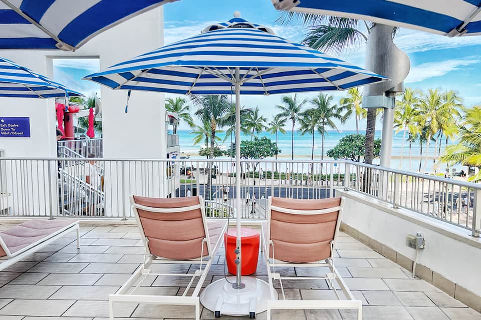 Beachfront terrace with blue-striped umbrellas, coral lounge chairs, ocean and palm trees visible