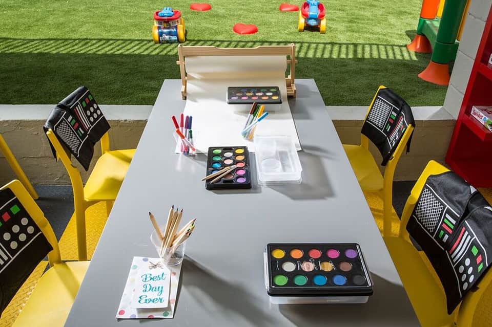 Children's activity table with art supplies, paint palettes, and colorful chairs overlooking outdoor play area