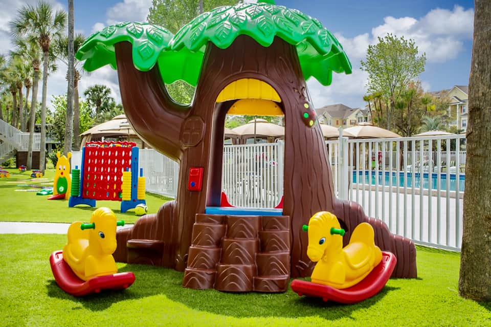 Colorful tree-shaped playground structure with yellow duck rockers on manicured lawn near residential community
