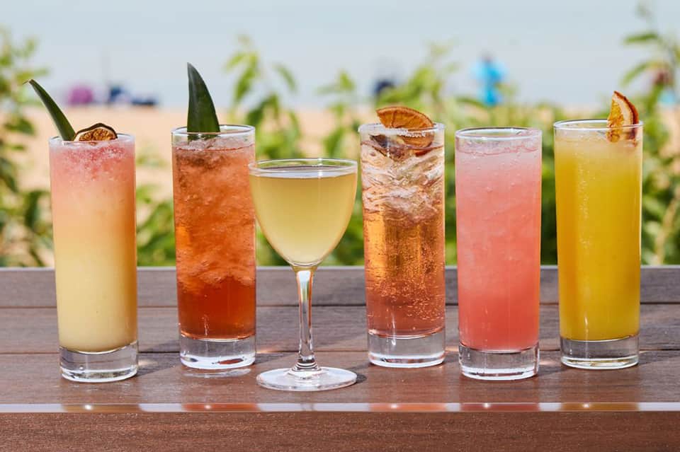 Six colorful tropical cocktails lined up on a wooden bar with beach background