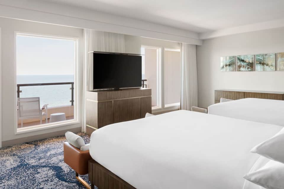 Modern oceanfront hotel bedroom with two queen beds, flat-screen TV, and sliding glass doors opening to a balcony overlooking the sea