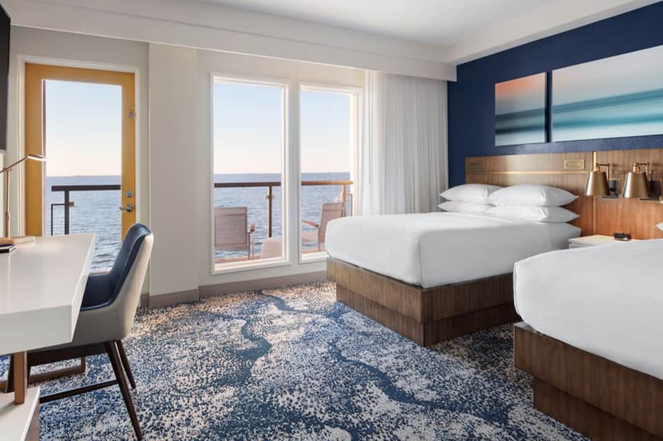 Modern hotel bedroom with ocean views, queen beds, desk, and glass balcony doors