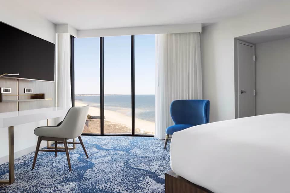 Beachfront hotel room with oceanview, white bed, blue accent chair, desk, and floor-to-ceiling windows