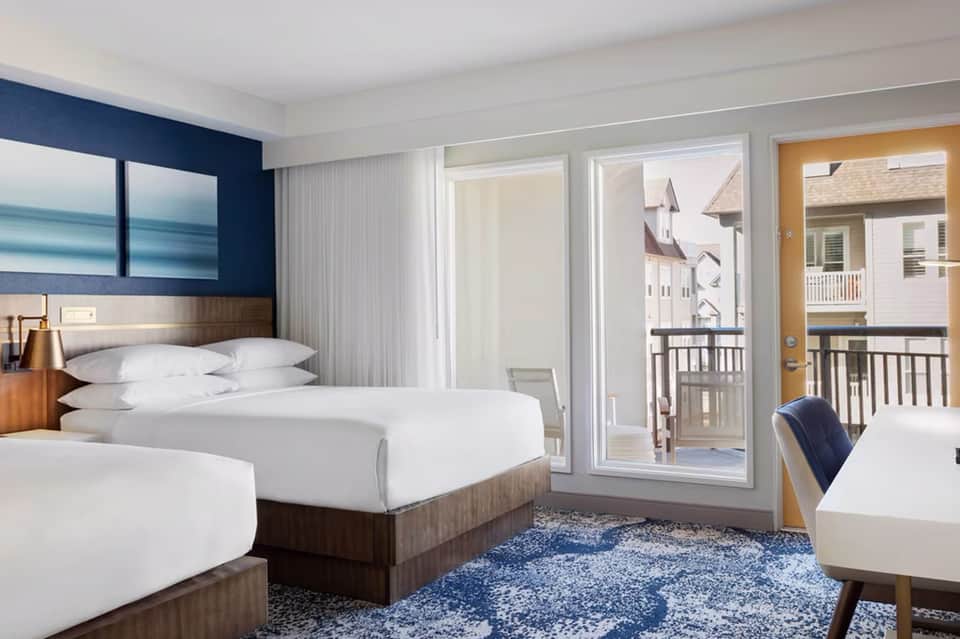 Modern hotel room with two beds, dark blue accent wall, ocean artwork, and balcony overlooking coastal homes