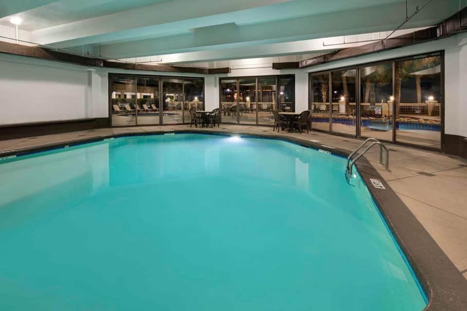 Indoor swimming pool with turquoise water, ceiling beams, lounge chairs, and glass doors to outdoor patio