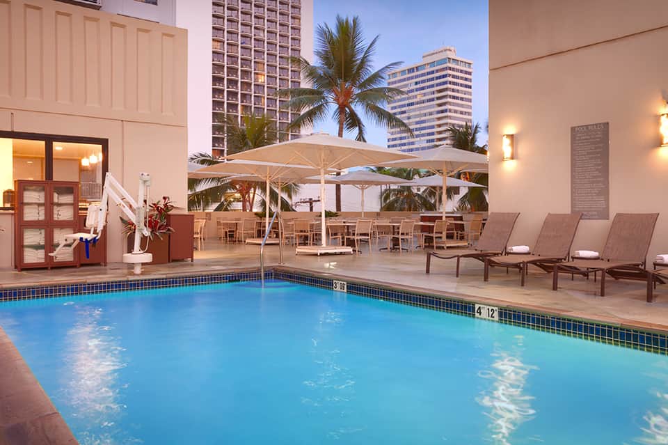 Resort pool with blue water, lounge chairs, umbrellas, and tall hotel buildings in tropical setting
