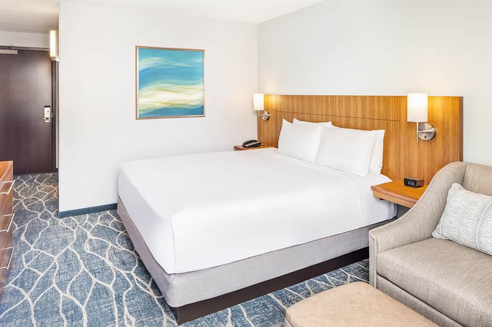 Modern hotel bedroom with queen bed, wood headboard, beige armchair, and abstract blue artwork