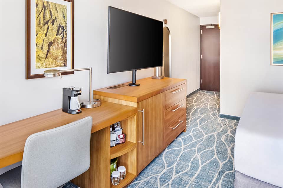 Modern hotel room with wooden desk, flat-screen TV, coffee maker, and patterned carpet flooring