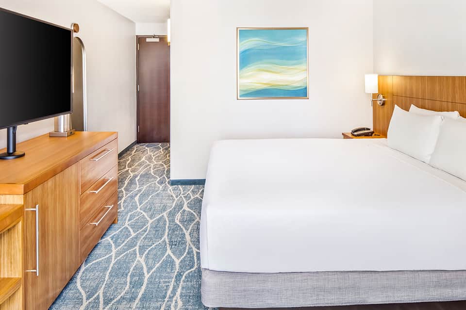 Modern hotel room with queen bed, wooden dresser, flat-screen TV, and ocean-themed artwork