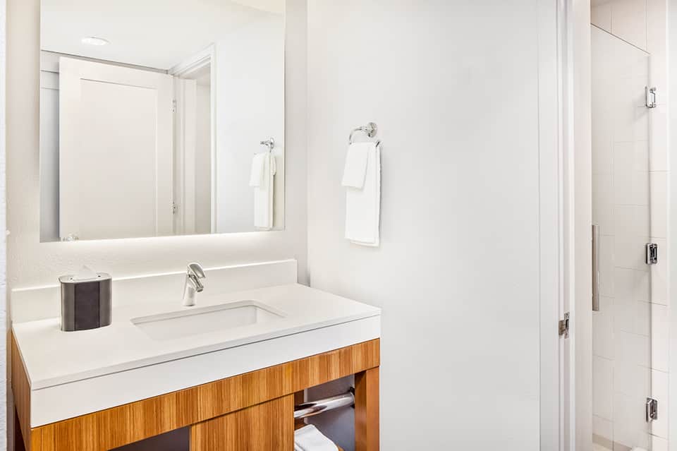 Clean minimalist bathroom with white vanity, wooden cabinet base, large mirror, and tiled shower area