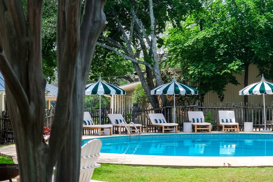 Resort pool with striped umbrellas and lounge chairs surrounded by mature trees and wooden deck