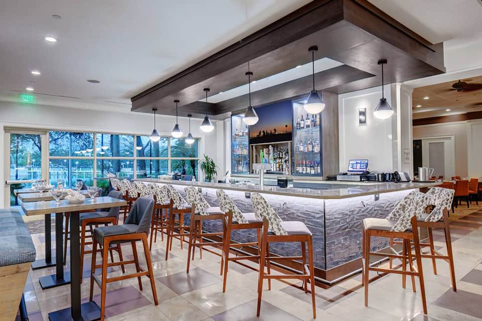 Modern hotel bar and lounge with pendant lights, marble counter, wood beam ceiling, and large windows overlooking trees