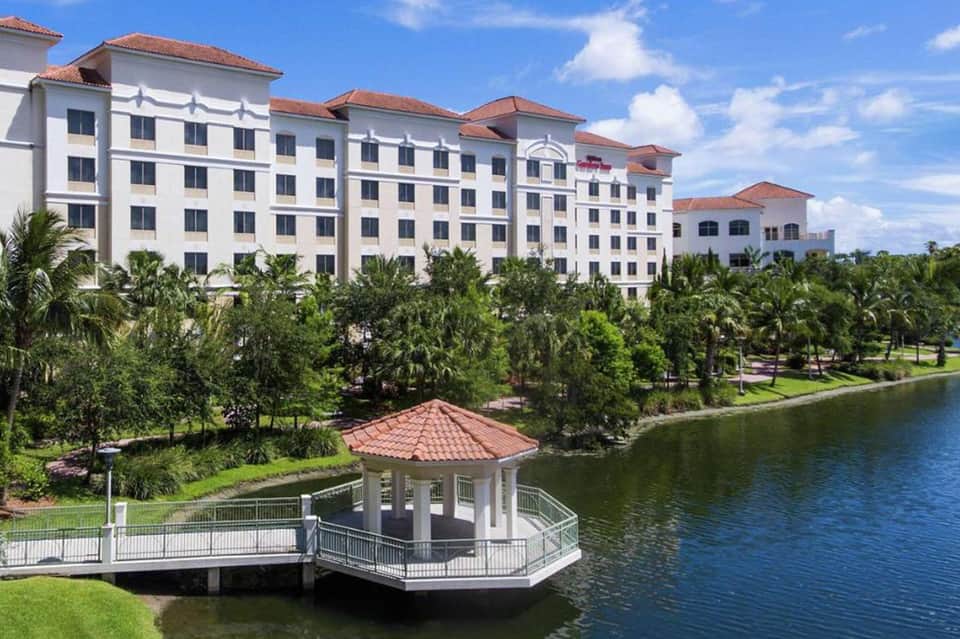 Waterfront hotel with white exterior, terracotta roof, surrounded by palm trees and manicured lawns overlooking a lake