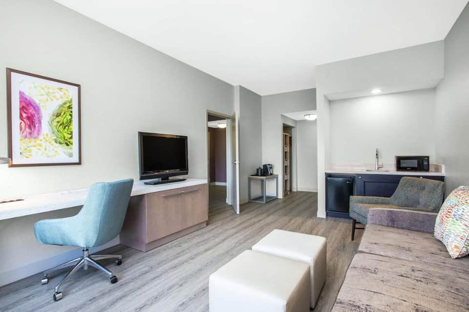 Modern hotel suite with desk, TV, seating area, and kitchenette with neutral tones