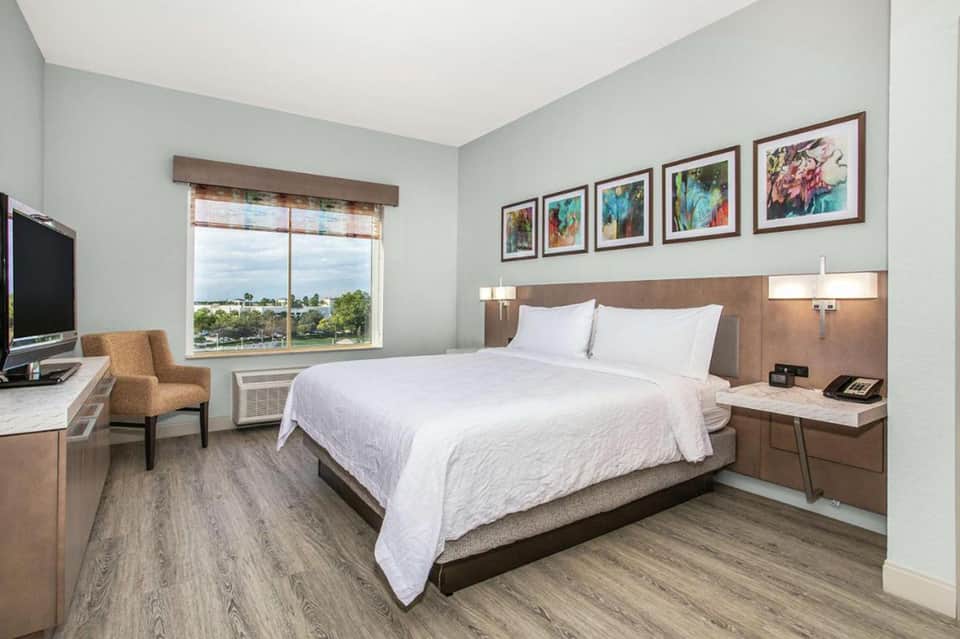 Hotel bedroom with queen bed, wood furnishings, TV, chair, and large window overlooking green landscape