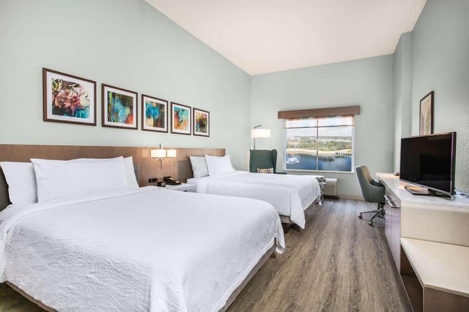 Bright hotel room with two queen beds, water view windows, framed artwork, desk area, and light wood flooring