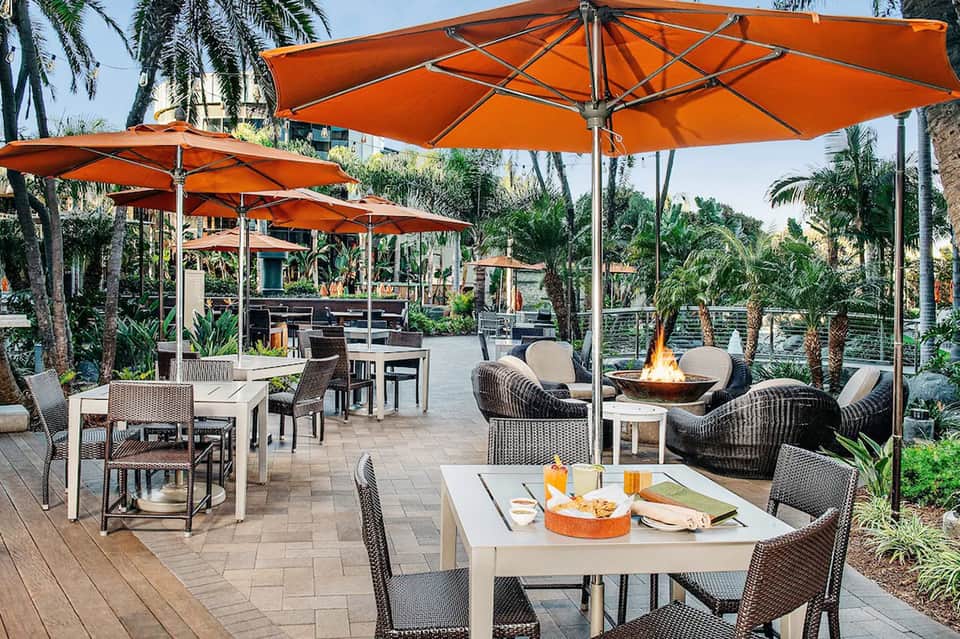 Outdoor patio with orange umbrellas, wicker seating, fire pit, and palm trees at dusk