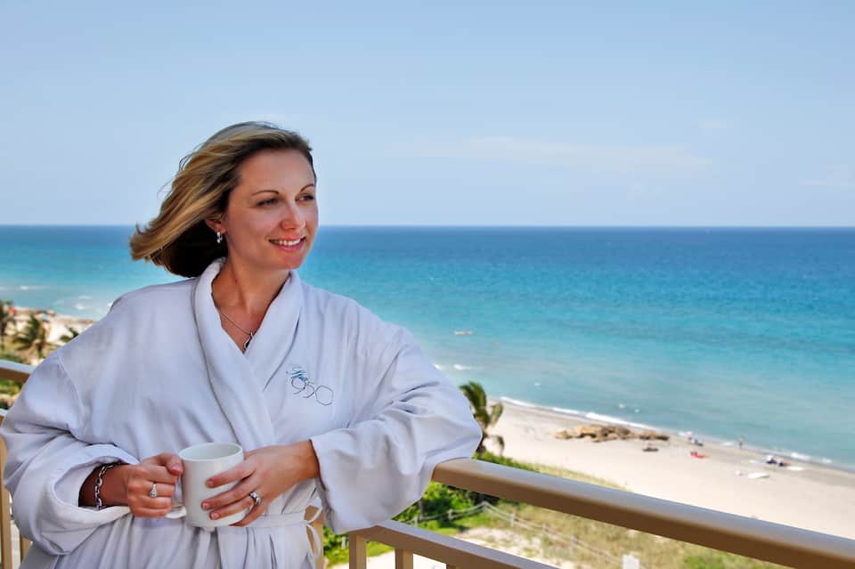 Woman in white spa robe holding coffee cup on oceanfront balcony overlooking beach
