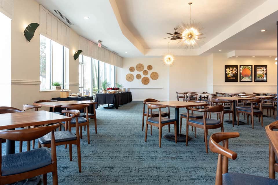 Modern hotel dining room with wooden tables, blue carpet, decorative woven wall plates, and large windows