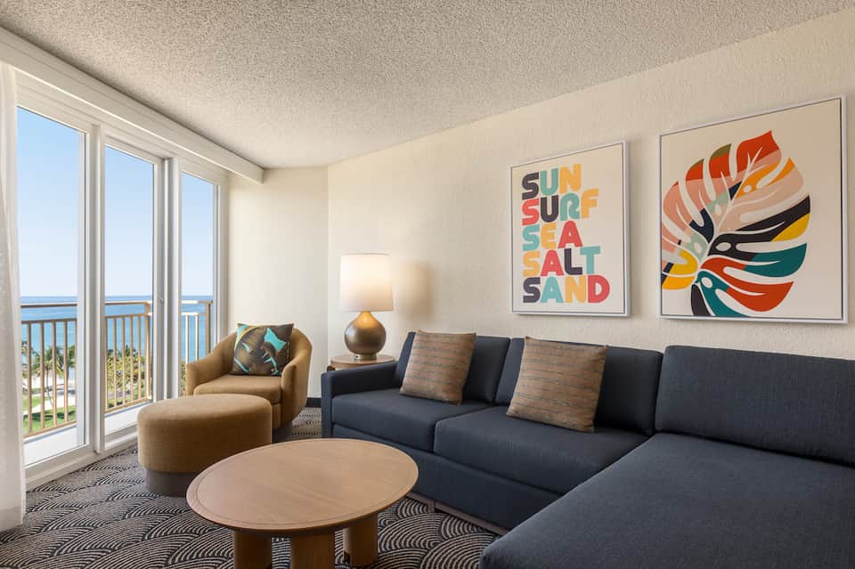 Coastal hotel suite with ocean view, modern sofa, colorful wall art, and balcony access