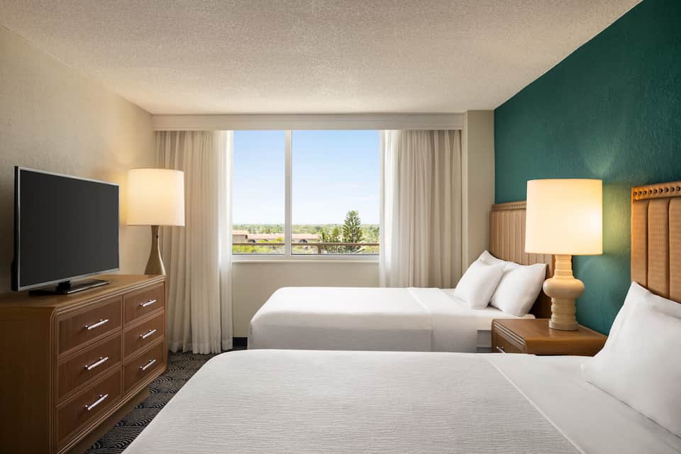 Hotel room with two queen beds, teal accent wall, wooden furniture, and large windows overlooking landscape